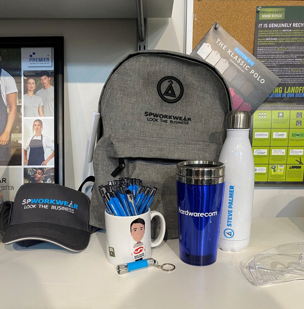 Assorted promotional items including embroidered caps, branded backpacks, printed mugs, water bottles, and keychain torches and pens from SP Workwear